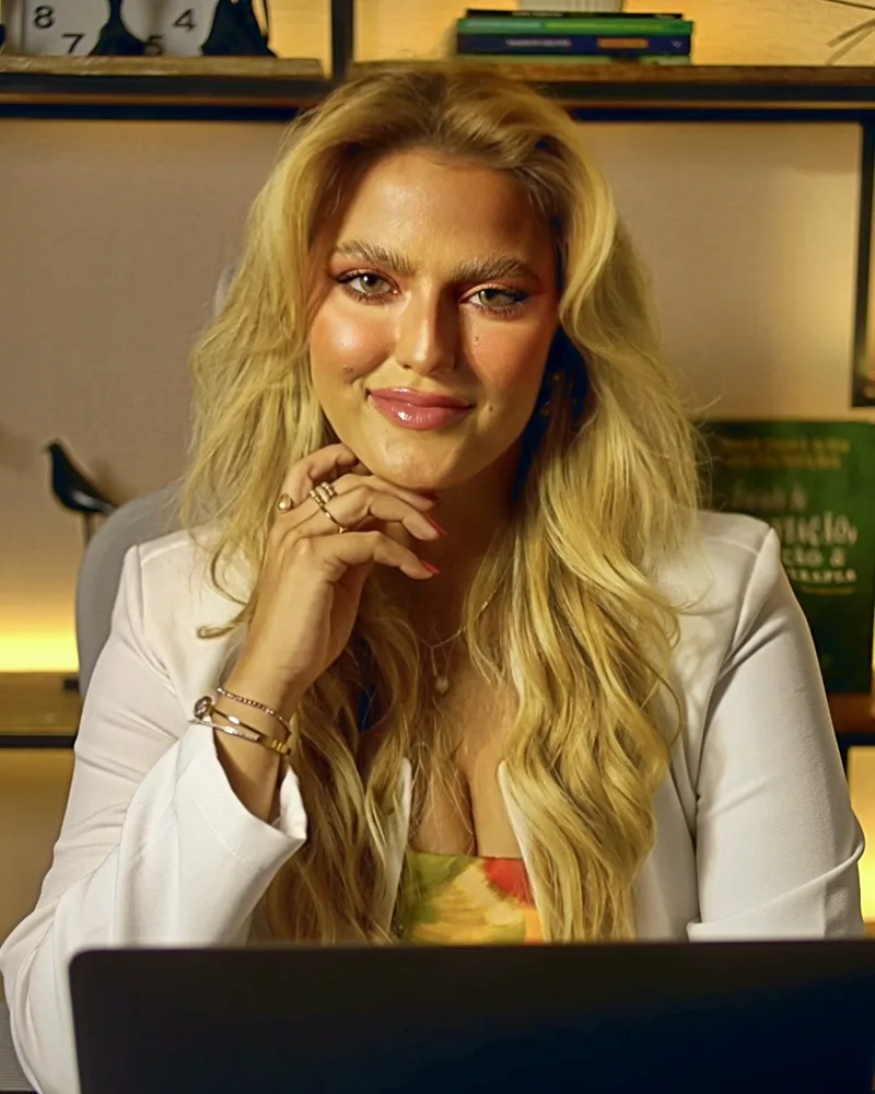 Fanny Viana sorrindo para a câmera, de blazer branco em seu escritório, com uma estante iluminada ao fundo